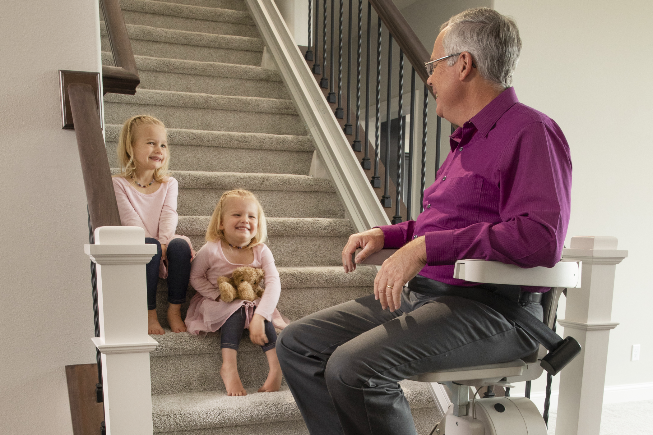 An older man riding a stairlift interacts with two children seated on carpeted stairs, with a stuffed bear beside them. The setting appears to be a home interior with wooden railing.