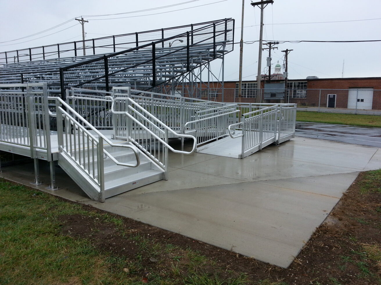 A set of metal bleachers with railed ramps and steps is situated on a concrete platform next to a grassy area on a rainy day. A brick building is visible in the background.