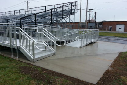 A set of metal bleachers with railed ramps and steps is situated on a concrete platform next to a grassy area on a rainy day. A brick building is visible in the background.