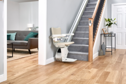 A beige stairlift installed on a carpeted staircase adjacent to a hallway with wooden flooring, leading to a living room with a grey sofa and turquoise pillows.