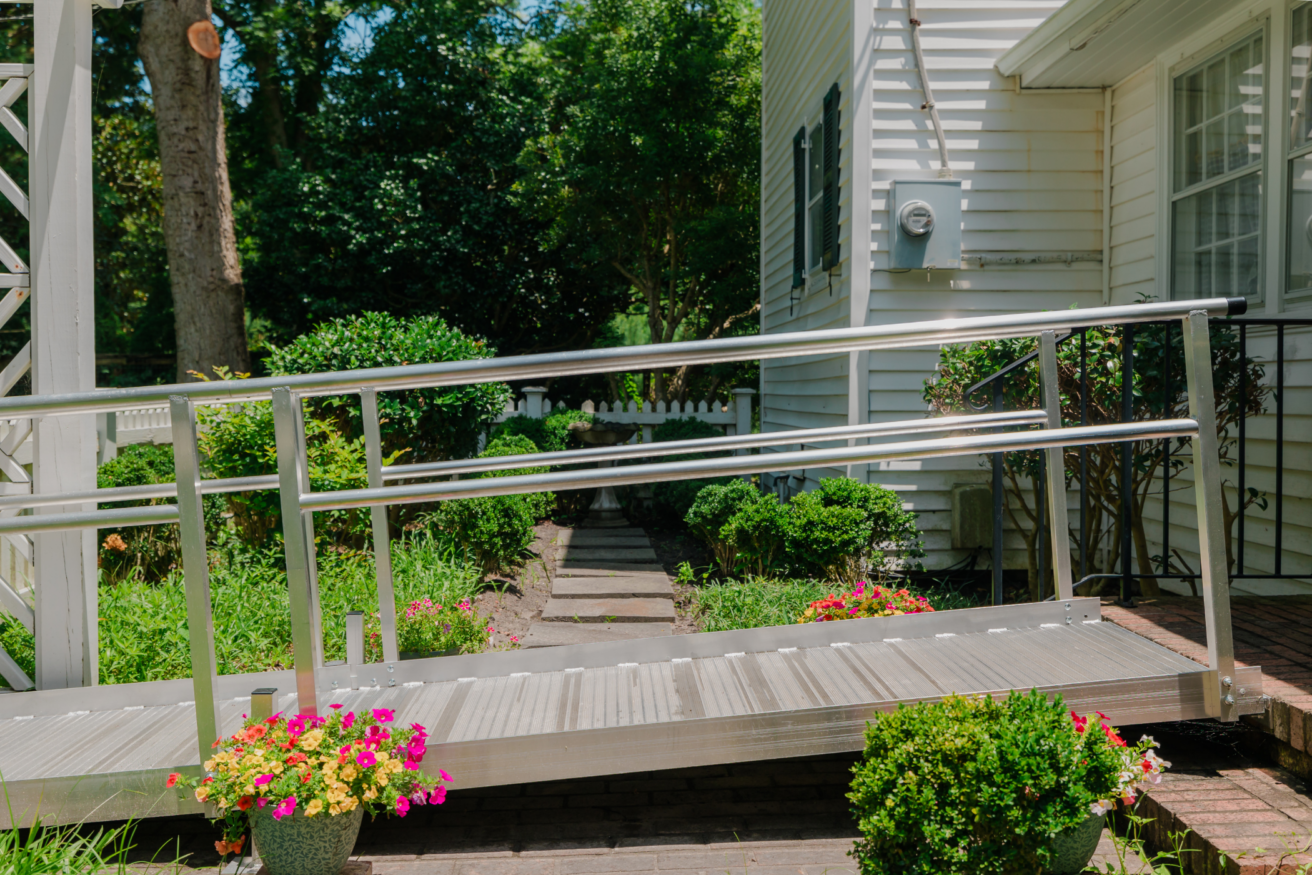 A metal wheelchair ramp leads to a house entrance. The yard has green shrubs, colorful flowers, and a paved walkway.