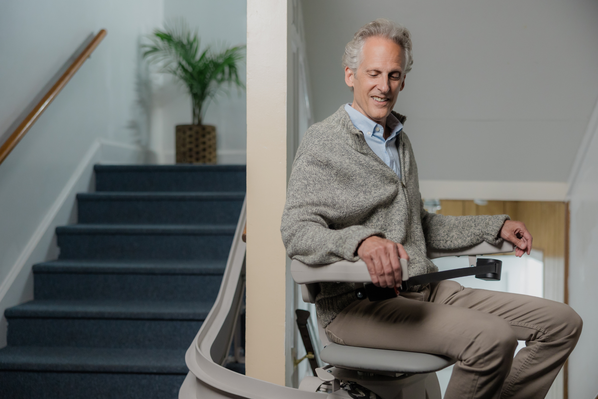 An older man with gray hair rides a stairlift, smiling and holding the armrest, next to a staircase in a home.