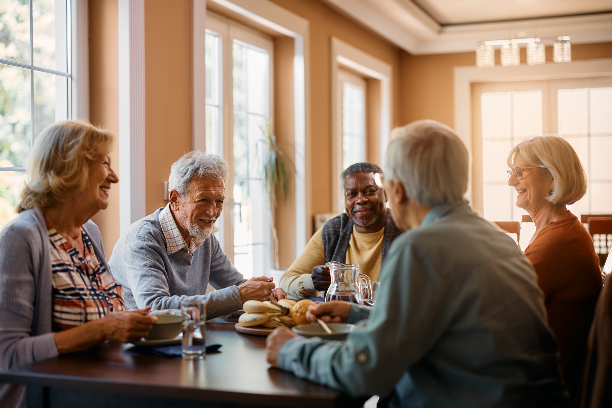 A group of five elderly people sit around a table in a well-lit room, engaging in conversation and sharing a meal.