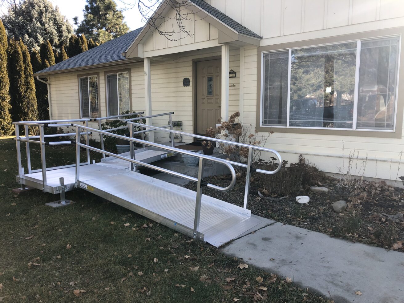 A single-story house with a wide, metal accessibility ramp leading to the front entrance. The ramp passes through a modest front yard with grass and some shrubs.