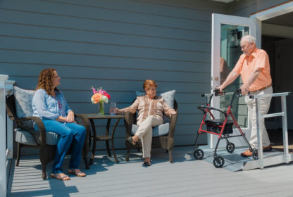 Three people are on a porch. One older woman and one younger woman sit on wicker chairs, while an older man stands using a walker. A small table with flowers is between the seated women.