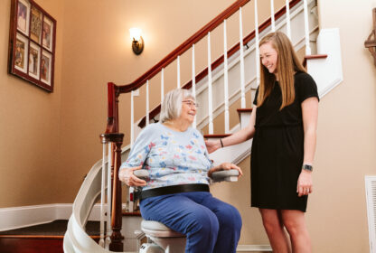 An elderly woman sits on a stairlift, smiling and talking to a young woman standing next to her in a home with beige walls and framed photos.