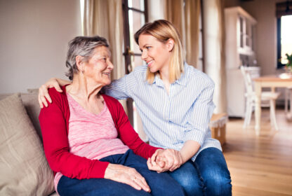 An elderly woman and a younger woman sit on a couch, smiling at each other. The older woman wears a red cardigan, while the younger woman wears a blue striped shirt and holds the older woman's hand.