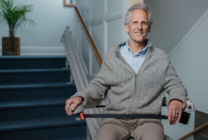 A man rides a stairlift in a residential setting, smiling at the camera. He is wearing a light gray sweater and khaki pants. There is a potted plant at the top of the staircase.