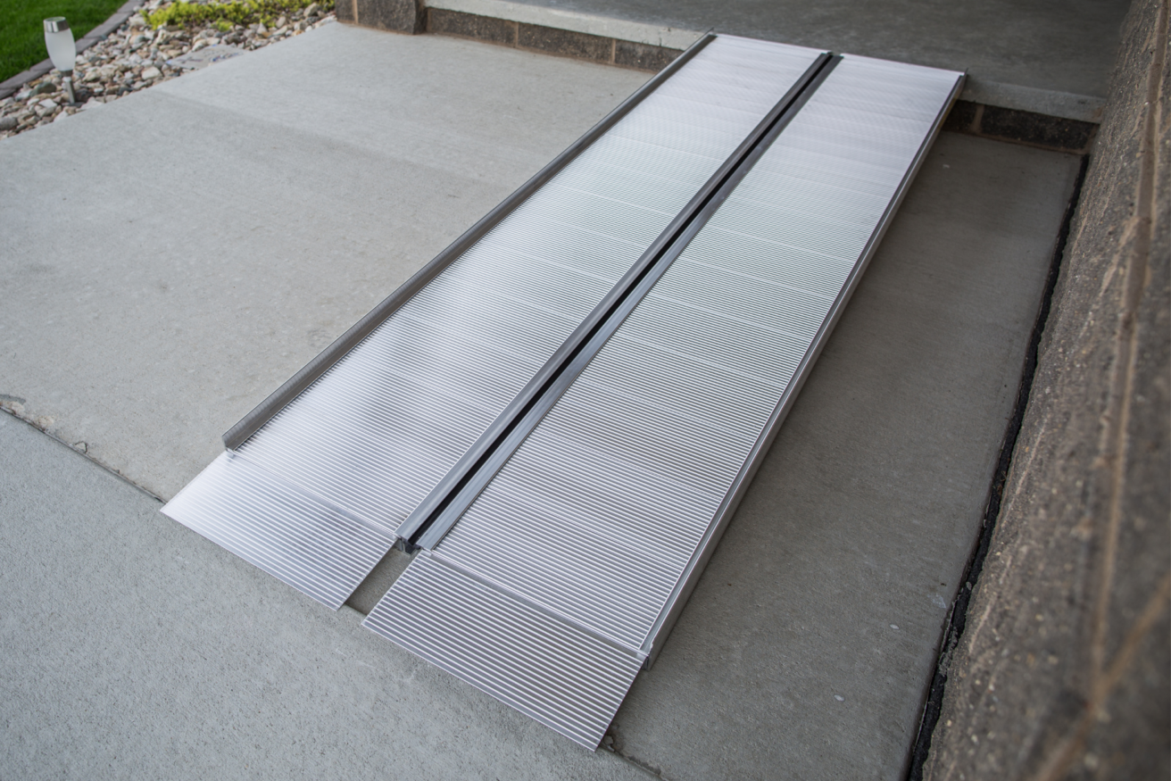 A metal wheelchair ramp placed over a step leading into a building. The ramp has a grooved texture for traction and extends from the ground to the doorway.