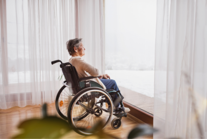 An elderly person sits in a wheelchair looking out of a large window with sheer white curtains, gazing at a snowy landscape outside.