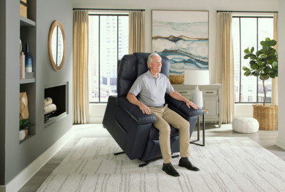 An elderly man sits in a blue recliner chair in a brightly lit living room with large windows, beige curtains, a wall mirror, and modern decor.