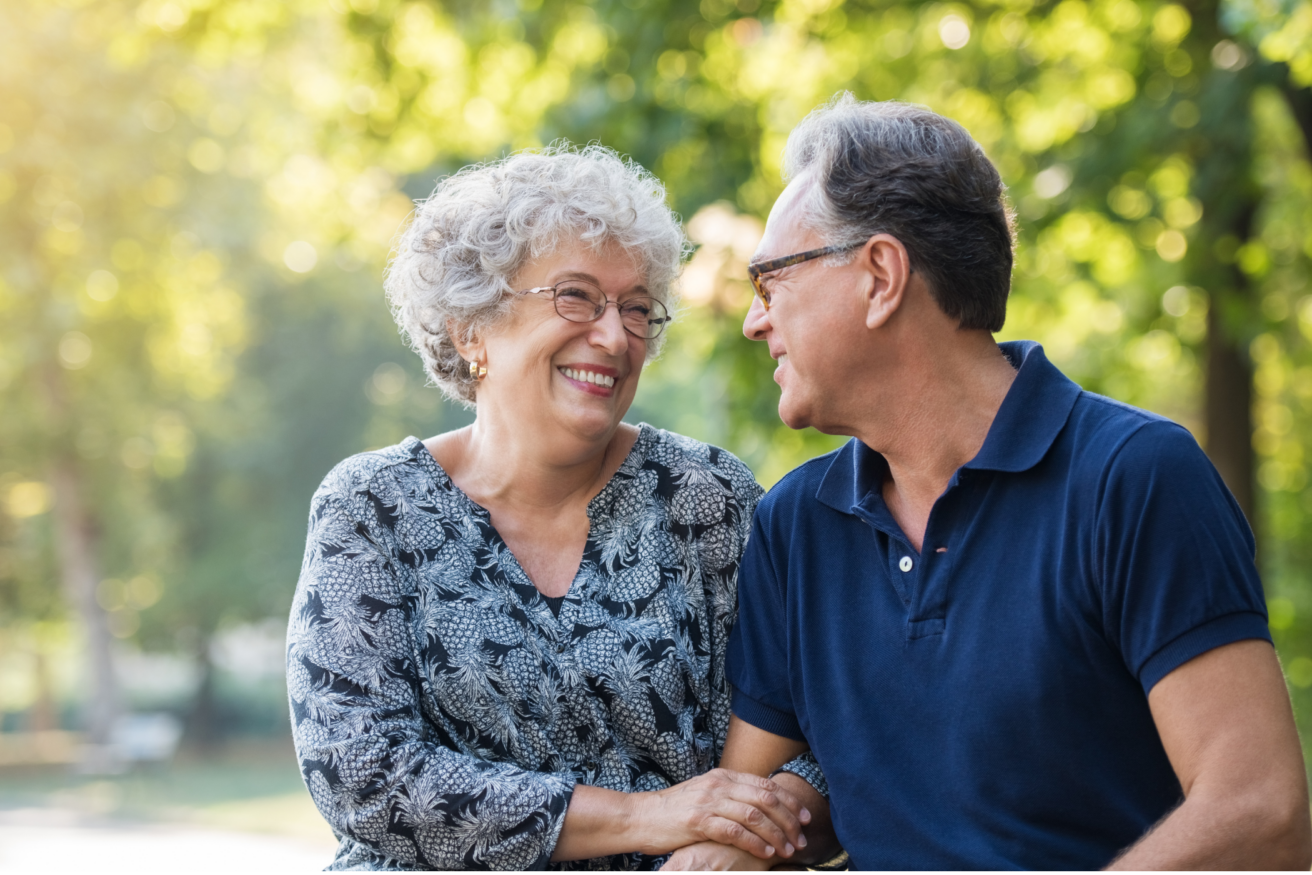 An elderly couple sits outdoors, smiling at each other. The woman wears glasses and a patterned blouse; the man wears glasses and a blue polo shirt. Trees and sunlight are in the background.