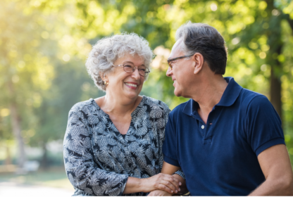 An elderly couple sits outdoors, smiling at each other. The woman wears glasses and a patterned blouse; the man wears glasses and a blue polo shirt. Trees and sunlight are in the background.