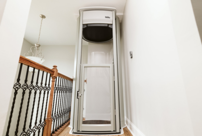 A small home elevator with a glass door and white frame installed on the floor next to a wooden railing and metal balusters in a hallway.