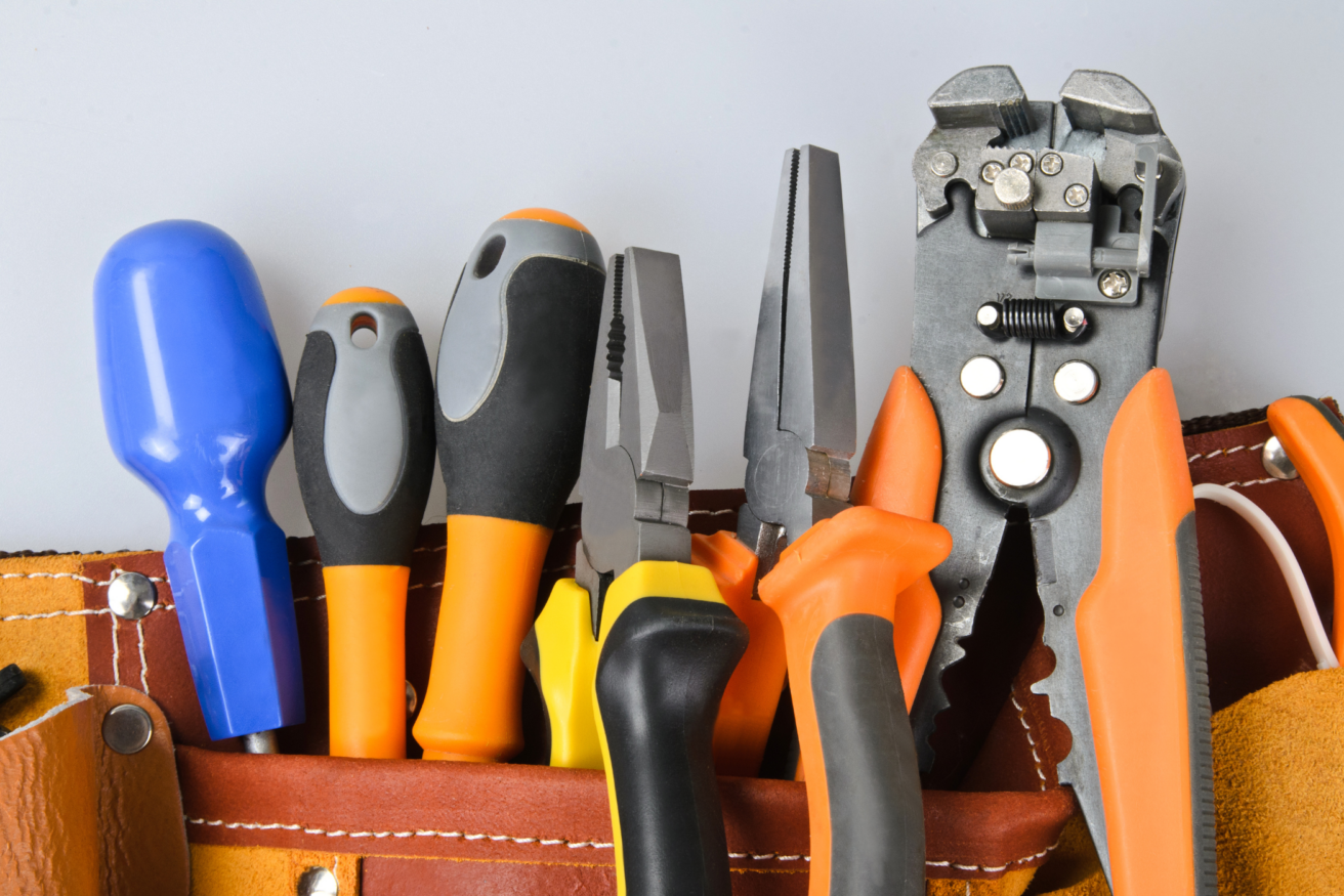 Close-up of various hand tools including screwdrivers, pliers, and wire strippers in a brown leather toolbelt against a light grey background.