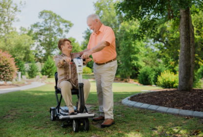 An elderly woman in a mobility scooter is being assisted by an elderly man in an outdoor park setting. Both are smiling.