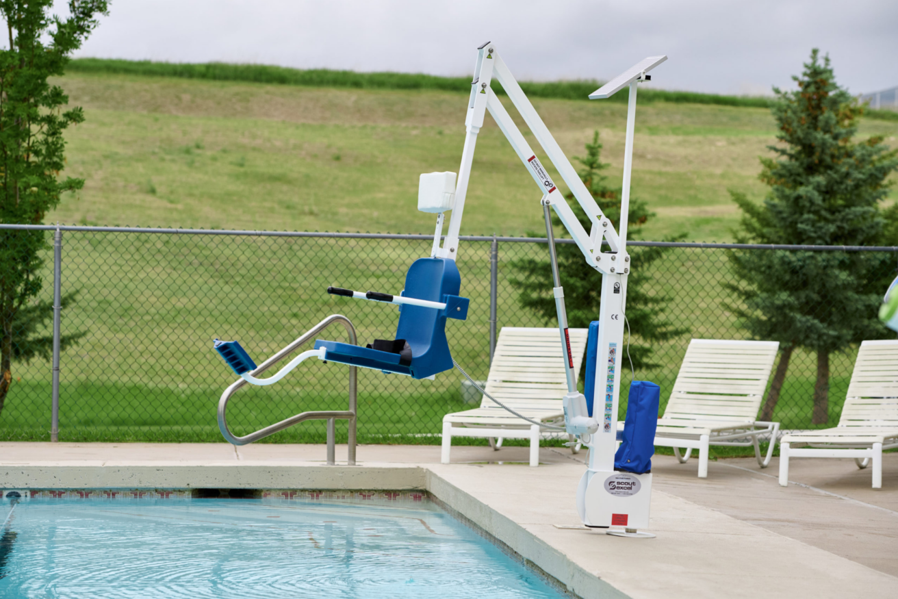 A pool lift at the edge of an outdoor swimming pool with lounge chairs and a green lawn in the background.