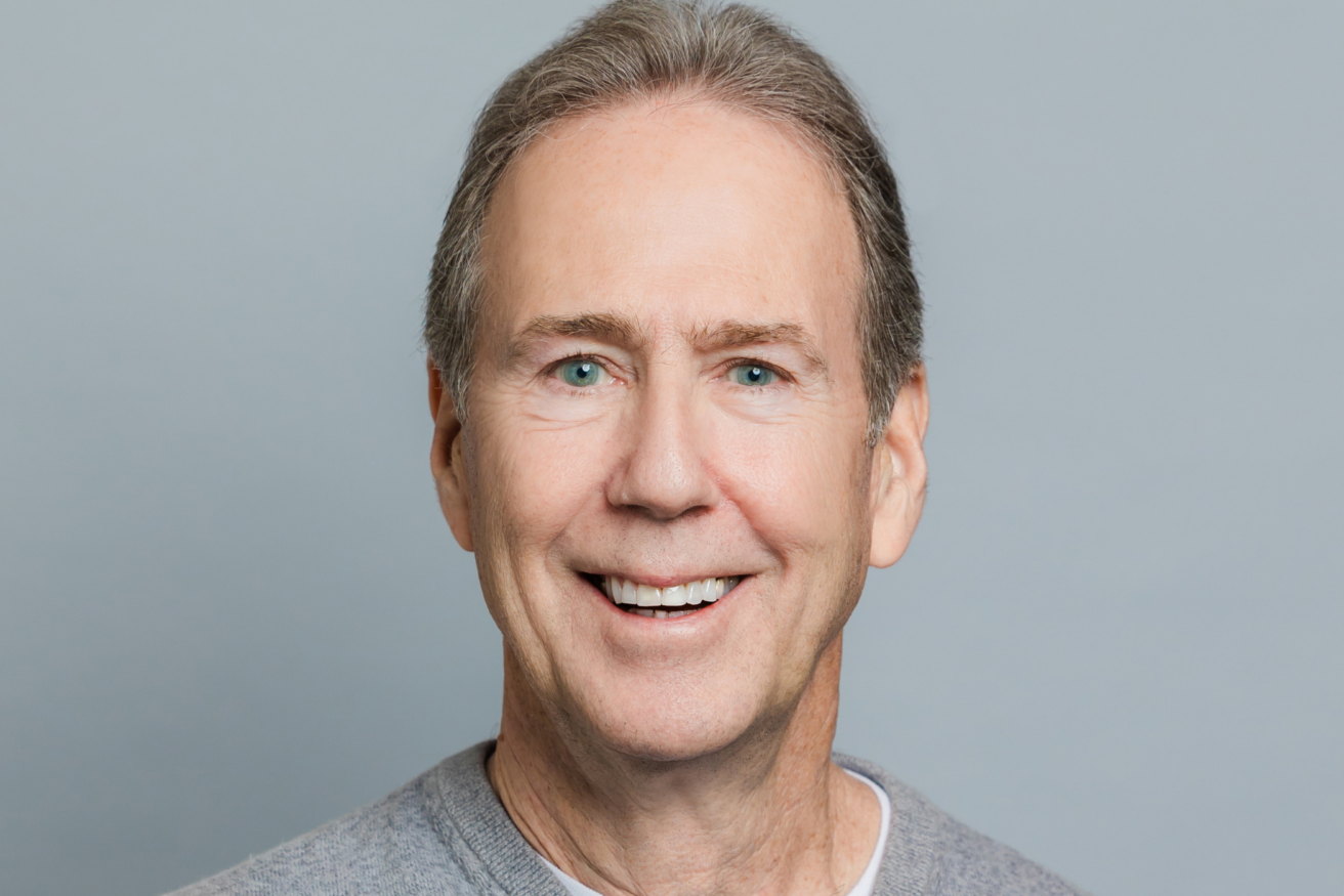 A man with short gray hair and wearing a gray sweater smiles at the camera against a plain gray background.