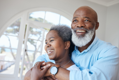 Two people smiling and embracing in a bright room with arched windows. The person in front has short gray hair, and the person behind has a gray beard and wears a light blue shirt.