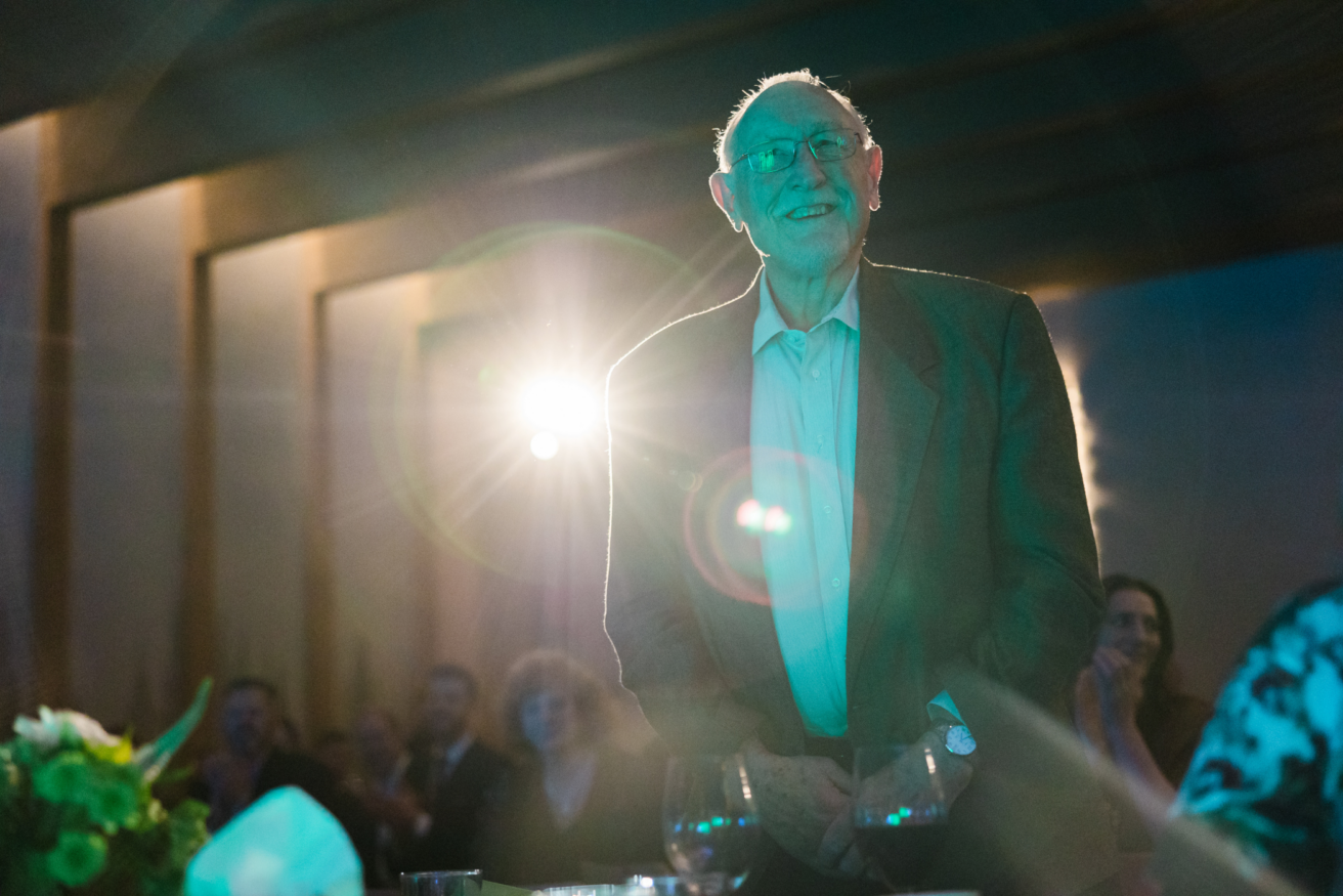 An elderly man in a suit stands and smiles at an event, with bright stage lighting in the background and a seated audience.