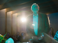 An elderly man in a suit stands and smiles at an event, with bright stage lighting in the background and a seated audience.