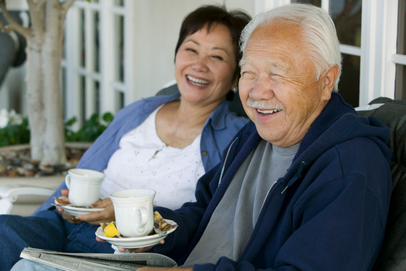 An elderly couple sits together on a porch, both smiling and holding teacups, with a newspaper in the man's lap.