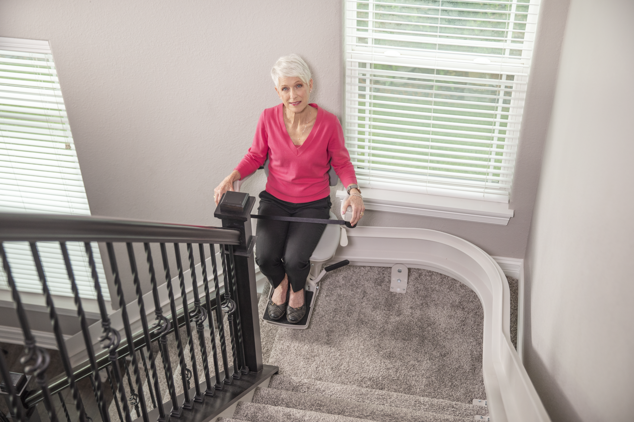 An elderly woman with short white hair sits on a stair lift chair at a corner bend in a carpeted staircase, holding onto the chair controls and looking at the camera.