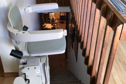 A chair lift is installed along the staircase of a home, providing accessibility between floors. The stair lift has a grey and white seat and runs parallel to the wooden handrail.