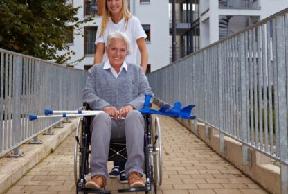 A person in a gray sweater sits in a wheelchair holding crutches, while another person in a white shirt stands behind, pushing the wheelchair along a paved path.