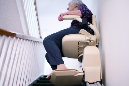 An elderly person sits on a stairlift, ascending a narrow staircase, with one hand resting on the armrest.