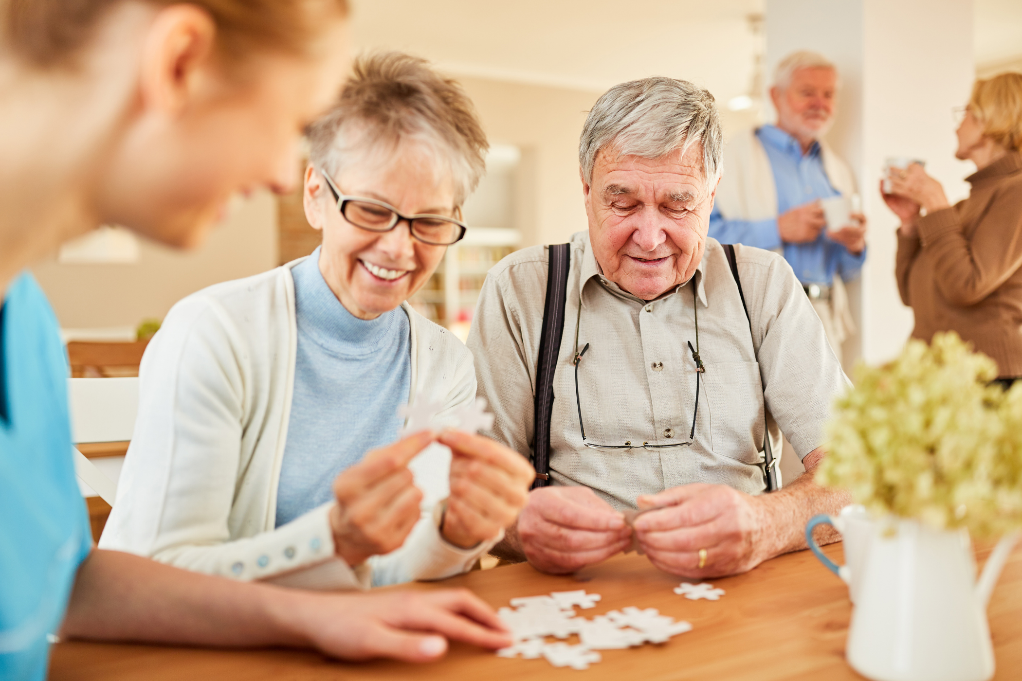 Two senior people assemble a puzzle at a table while a younger person assists. In the background, another senior man and woman engage in conversation and hold drinks.