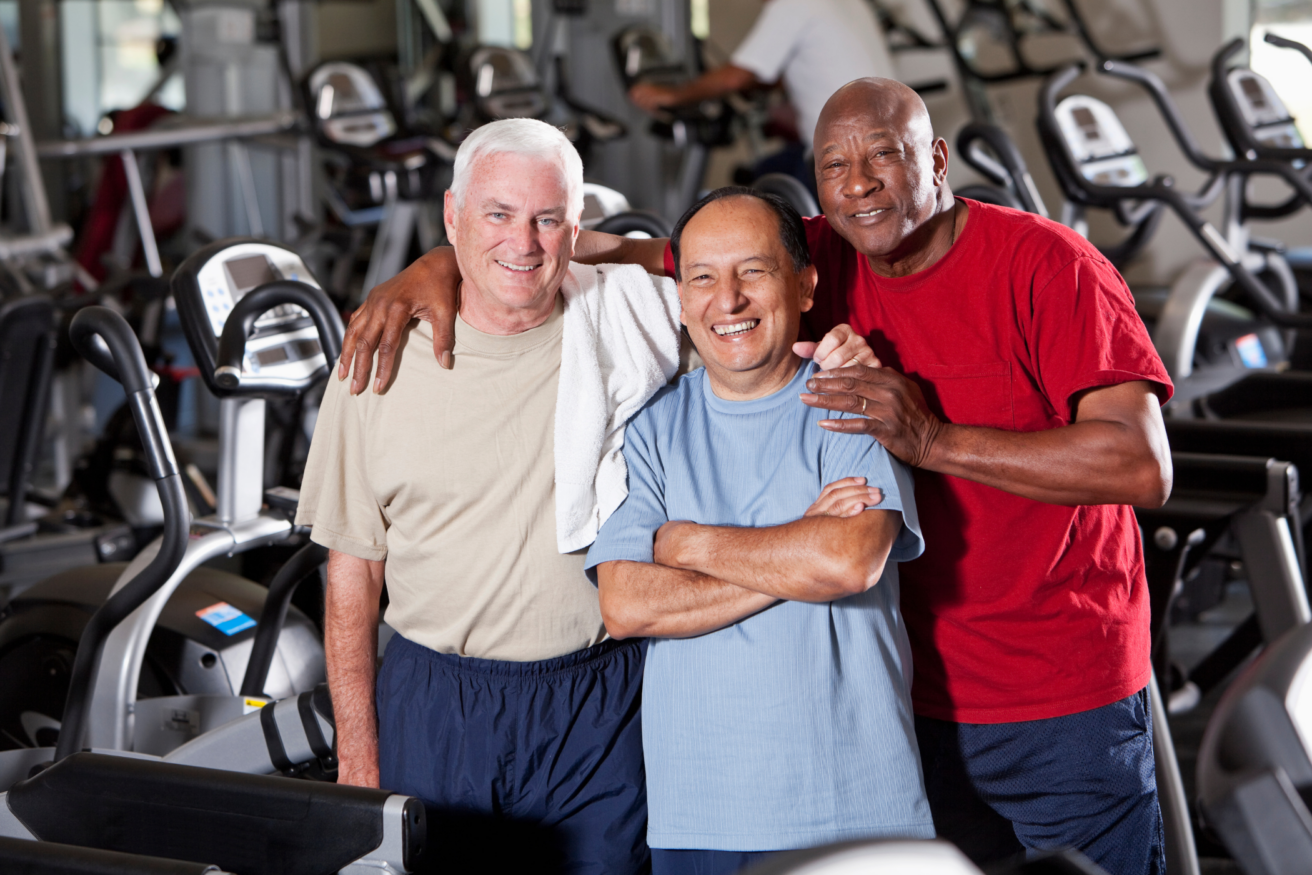 Three men standing together in a gym, smiling, with exercise equipment in the background. One man has a towel draped over his shoulder.