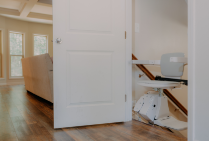 A stair lift is installed on a wooden staircase near an open door in a well-lit room with a beige sofa and windows in the background.