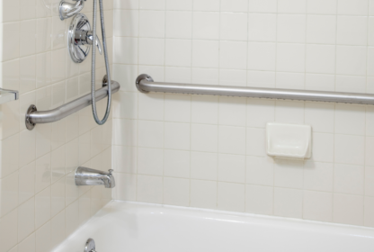 A bathroom with a white tiled wall featuring a bathtub, a metal handrail, a wall-mounted showerhead, and a soap dish.