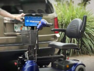 A person holds a Harmar lift attached to a vehicle with a wheelchair secured on it. The vehicle's license plate reads "WHY" from Florida. The scene includes greenery in the background.