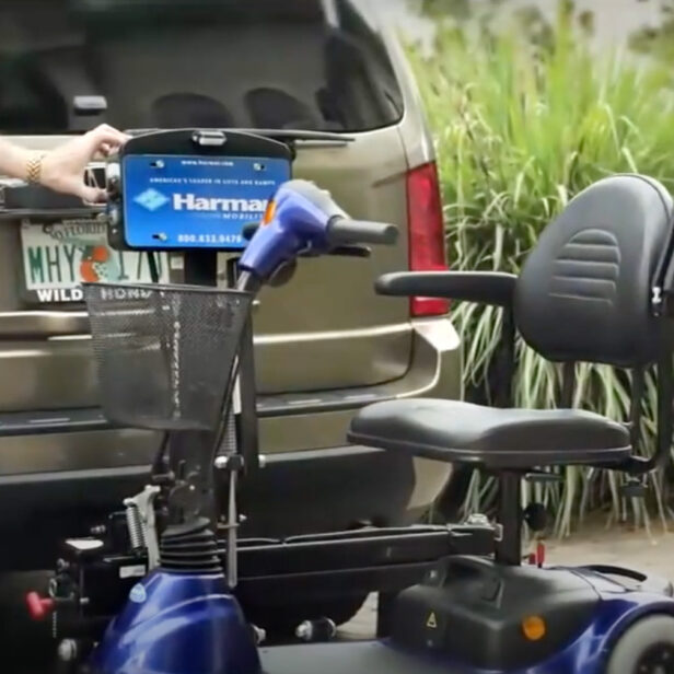 A person holds a Harmar lift attached to a vehicle with a wheelchair secured on it. The vehicle's license plate reads "WHY" from Florida. The scene includes greenery in the background.