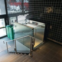 A modern, indoor wheelchair lift with glass panels adjacent to a staircase. The background shows a black tiled wall and green recycling bins near an entrance door.