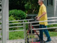 An older man wearing a yellow shirt and blue jeans uses a walker to navigate a wheelchair ramp outside a house with a garden.