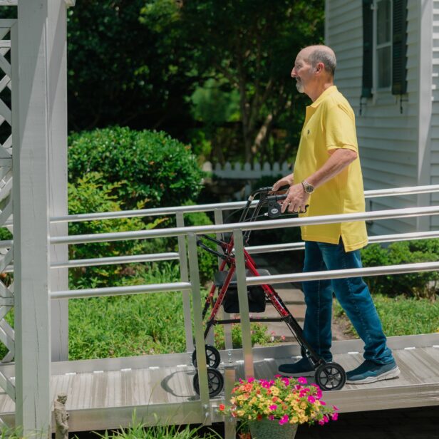 An older man wearing a yellow shirt and blue jeans uses a walker to navigate a wheelchair ramp outside a house with a garden.