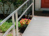 A metal accessibility ramp leads to a house with an orange door. Flowers and greenery are visible beside the ramp.