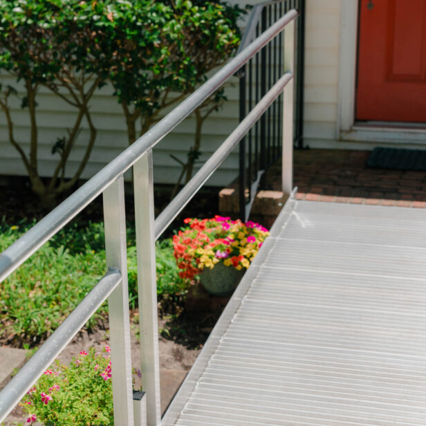 A metal accessibility ramp leads to a house with an orange door. Flowers and greenery are visible beside the ramp.