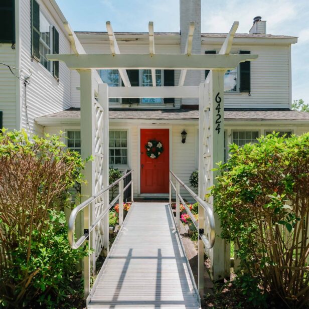 A white house with a red door and a wreath. A ramp leads to the entrance under a white pergola labeled "6424." Green bushes line the pathway.