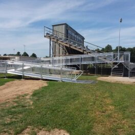 An outdoor stadium structure with a metal ramp leading up to tiered seating and a press box. The grassy area around the ramp shows patches of dirt.