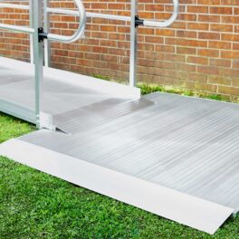 A metal wheelchair ramp with railings leads up to a platform, set against a brick wall and grass-covered ground.