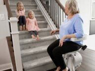 An elderly woman is using a stair lift while two young children sit on the carpeted stairs, smiling and interacting with her.