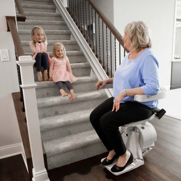 An elderly woman is using a stair lift while two young children sit on the carpeted stairs, smiling and interacting with her.