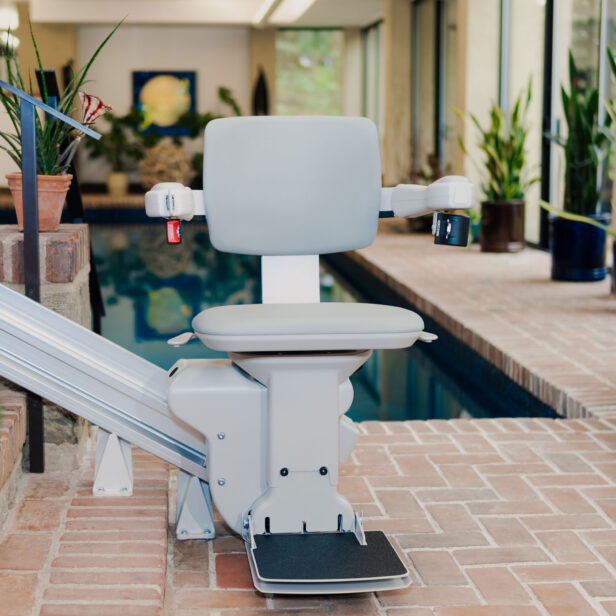 A gray stair lift chair is installed next to an indoor pool, surrounded by brick flooring and potted plants in a brightly lit room.