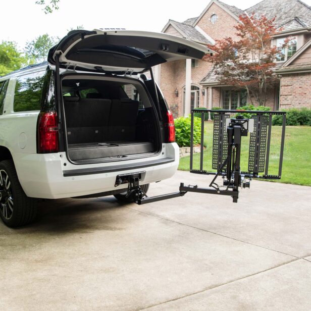 A white SUV with an open trunk is parked in a driveway. A foldable platform lift is attached to the rear hitch of the SUV, extending out towards the ground. A brick house is in the background.