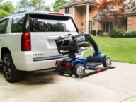 A white SUV is parked in a driveway with a folded mobility scooter attached to the rear on a carrier lift. A brick house with a well-maintained lawn is visible in the background.