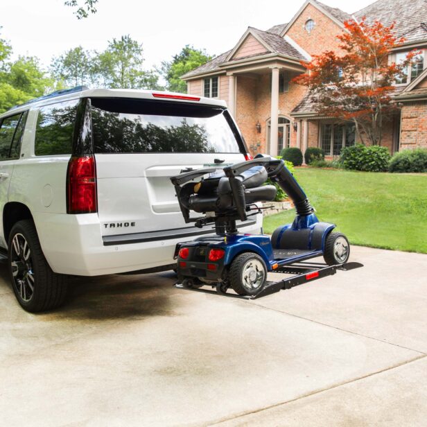 A white SUV is parked in a driveway with a folded mobility scooter attached to the rear on a carrier lift. A brick house with a well-maintained lawn is visible in the background.